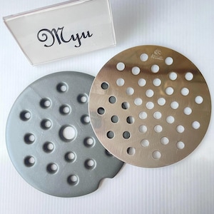 May include: A gray circular silicone mat with a perforated pattern and a metallic circular drain cover with a matching hole pattern. A clear acrylic sign with the text Myu is positioned above the two circular items.
