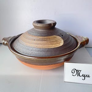 以下が含まれることがあります： 濃い茶色のテクスチャーのある蓋にベージュの水平な帯が入った、丸い陶器製の土鍋。本体は滑らかな赤茶色の粘土で、光沢のある仕上げが施されています。両側に持ち手があり、黒い文字でMyuと書かれた白い看板が添えられています。