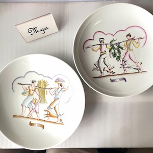 以下が含まれることがあります： 農業シーンの人物が描かれたカラフルなイラスト入りの白い陶器製プレート2枚。1枚には「ESTATE」の文字と共に収穫の様子が、もう1枚には「AVTVNNO」の文字と共にブドウ収穫の様子が描かれています。「Myu」と書かれた小さなカードが添えられています。