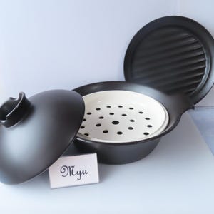 以下が含まれることがあります： 黒いセラミック製の蒸し器セット。丸いベース、白い穴あきインサート、黒い蓋、黒い溝付きプレートで構成されています。セットの前にはMyuという文字が書かれた小さな白い長方形の看板が置かれています。