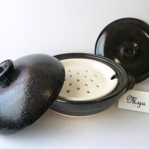 以下が含まれることがあります： 黒い釉薬をかけたセラミック製の調理鍋で、お揃いの蓋と、複数の円形の穴が開いた取り外し可能な白い蒸し器用トレイが付いています。Myuという文字が書かれた小さな白い長方形のカードが、調理容器の横に置かれています。