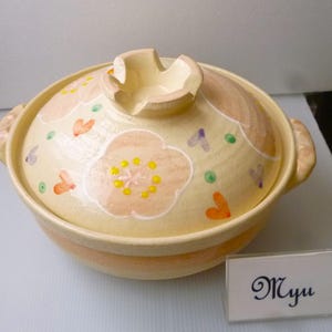 以下が含まれることがあります： クリーム色の陶器製土鍋。蓋には手描きの淡いピンクの花、黄色の中心部、オレンジと紫の小さなハート型のアクセントが施されています。蓋のつまみは切り込みが入った形状です。横には「Myu」と書かれた小さなプレートが置かれています。