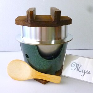 May include: A kamameshi rice cooker set featuring a dark green metal stove base, a silver aluminum pot, and a wooden lid with a handle. A light-colored wooden rice paddle is placed in front, alongside a small sign with the text Myu.