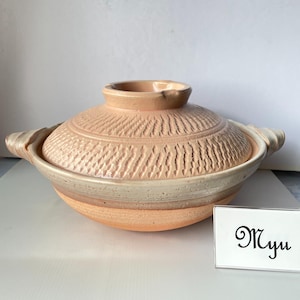 以下が含まれることがあります： 蓋付きの桃色の陶器製鍋。蓋には彫り込まれたテクスチャ模様があり、両側に持ち手が付いています。鍋の上部には滑らかな明るい色の帯状の装飾があります。小さな白い看板にはMyuという文字が書かれています。