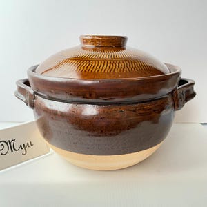 May include: A round, brown glazed ceramic pot with a matching lid featuring a textured, radial pattern. The pot has two side handles and a smooth, light-colored base. A white card with the text Myu is positioned next to the ceramic vessel.
