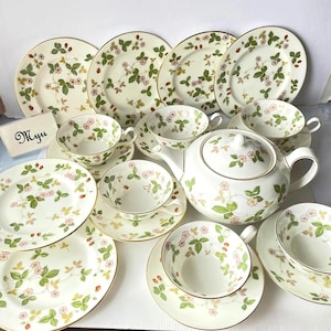 May include: A white porcelain tea set featuring a repeating pattern of green leaves, pink flowers, and red strawberries. The set includes multiple plates, teacups with matching saucers, and a teapot with a lid. A small card displays the text Myu.