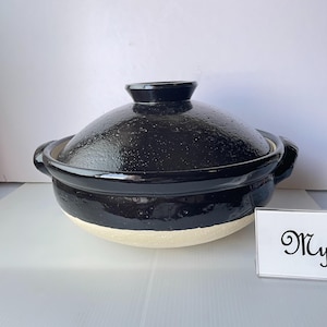 Könnte beinhalten: Ein schwarzer Donabe-Topf aus Keramik mit passendem Deckel, der eine glänzende Oberfläche im oberen Bereich und eine strukturierte, matte cremefarbene Basis aufweist. Der Topf hat zwei Seitengriffe und einen Deckelknauf. Ein weißes Schild zeigt den Text My.