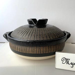May include: A round ceramic lidded pot with a textured dark brown and black finish. The lid features a radial grooved pattern and a central handle. The base is a light cream color. A small sign with the partial text My is visible.