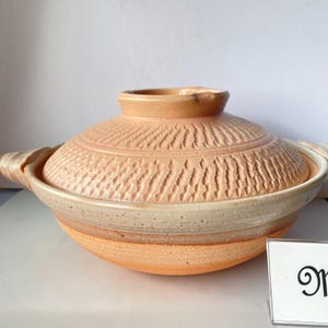 以下が含まれることがあります： 丸い薄オレンジ色の陶器製調理鍋と蓋のセット。蓋には縦線の彫り模様が施されています。鍋の上部には滑らかなクリーム色の帯があり、両側に持ち手が付いています。