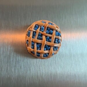 May include: A round, miniature pie-shaped magnet featuring a golden-brown lattice crust top. The filling consists of numerous small, round, bright blue berries visible through the gaps in the woven pastry strips. The object has a smooth, glossy finish.