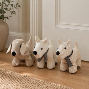 May include: Three fabric dog figurines standing in a row. The first is beige with white stars, the second is cream with a dark grey scarf, and the third is light tan with white hearts and a black and white checkered scarf. Each has black eyes.