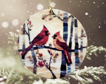 May include: A round, white ornament with a winter scene featuring two red cardinals perched on a branch. The background is a stylized depiction of birch trees with a snowy, wintery feel.