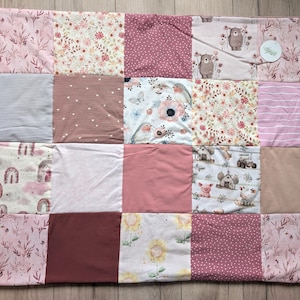 Könnte beinhalten: Eine Patchwork-Decke bestehend aus zwanzig quadratischen Feldern in verschiedenen Rosa-, Mauve- und Cremetönen. Die Stoffmuster zeigen Blumen, Streifen, Punkte, Regenbögen, Bären, Vögel, Sonnenblumen und Bauernhoftiere mit einem kleinen runden Etikett.