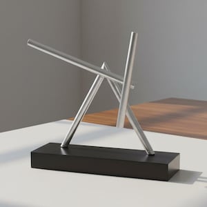 Puede incluir: Una escultura cinética que presenta tres varillas de metal de color plateado dispuestas en una formación angular e intersectada. Las varillas están sostenidas por una base rectangular de color negro sólido, creando una estructura equilibrada.