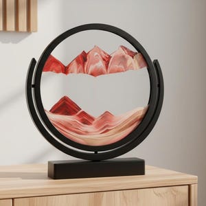 Op de afbeelding: Een cirkelvormig zwart frame gemonteerd op een rechthoekige zwarte voet met lagen rood, roze en wit zand. Het zand is gerangschikt om bergachtige pieken te vormen binnen de glazen behuizing, wat een gelaagd landschapseffect in de ring creëert.
