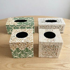 Puede incluir: Cuatro fundas para cajas de pañuelos rectangulares y cuadradas con incrustaciones de mosaico de conchas. Dos fundas muestran patrones de hojas y flores verdes, mientras que las otras dos presentan fragmentos de conchas blancas e iridiscentes.