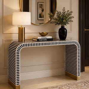 Può includere: Un tavolo console con una silhouette curva a cascata caratterizzata da un motivo a strisce bianche e nere e piastre di base in metallo dorato. La superficie del tavolo ospita una lampada dorata, una ciotola dorata, due libri impilati Chanel e Tom Ford e un vaso.