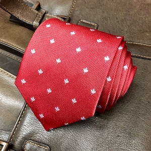 Op de afbeelding: Een opgerolde rode stropdas met een herhalend patroon van kleine witte esdoornbladontwerpen. De stropdas is gemaakt van een textuurstof en wordt weergegeven op een leren oppervlak.