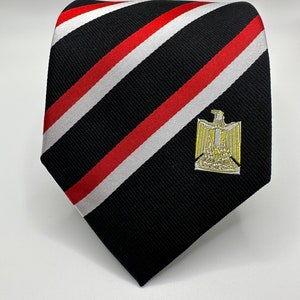 Op de afbeelding: Een zwart-rode gestreepte stropdas met een wit geborduurd adelaarsembleem. De adelaar heeft een gouden kroon en een gele snavel.
