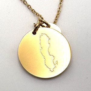 May include: A gold necklace with a round pendant featuring an outline of the shape of Sweden. The pendant is attached to a delicate gold chain.