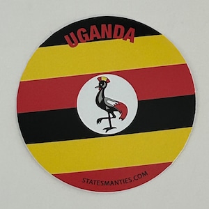 May include: Circular coaster displaying the Ugandan flag, with yellow, black, and red stripes. A central white circle contains a bird illustration. The word "UGANDA" is printed at the top in red.