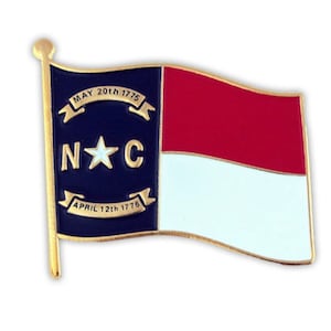 Puede incluir: Un pin de color dorado con la bandera del estado de Carolina del Norte. La bandera es azul con una estrella blanca en el centro y las letras "N*C" arriba y abajo de la estrella. La bandera est&aacute; rodeada por un borde dorado y tiene un poste dorado. La bandera ondea al viento. El texto en la bandera dice "MAY 20th 1775" y "APRIL 12th 1776".
