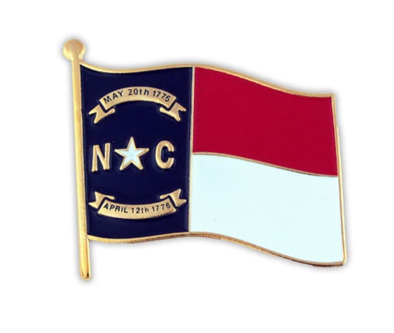 Puede incluir: Un pin de color dorado con la bandera del estado de Carolina del Norte. La bandera es azul con una estrella blanca en el centro y las letras "N*C" arriba y abajo de la estrella. La bandera est&aacute; rodeada por un borde dorado y tiene un poste dorado. La bandera ondea al viento. El texto en la bandera dice "MAY 20th 1775" y "APRIL 12th 1776".