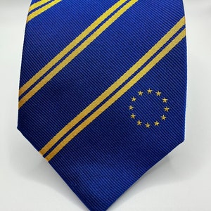 Op de afbeelding: Een blauwe zijden stropdas met diagonale gele strepen en een kleine geborduurde cirkel met gouden sterren in het midden.