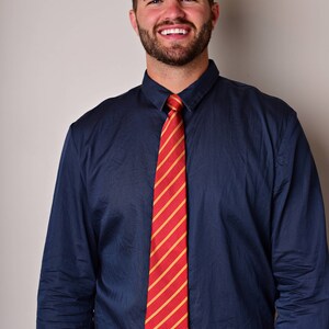 May include: A man wearing a navy blue dress shirt with a red and gold striped tie. He is smiling and wearing a brown leather belt with a silver buckle.