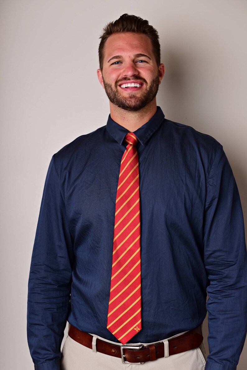 May include: A man wearing a navy blue dress shirt with a red and gold striped tie. He is smiling and wearing a brown leather belt with a silver buckle.