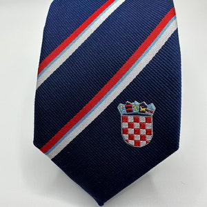 Op de afbeelding: Marineblauwe stropdas met diagonale strepen in rood, wit en lichtblauw. Het heeft een geborduurd embleem van een geblokt schild met een kroon. Een formeel accessoire voor zaken of speciale gelegenheden.