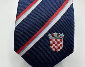 Może przedstawiać: Granatowy krawat z ukośnymi paskami w kolorze czerwonym, białym i jasnoniebieskim. Posiada haftowany herb w szachownicę z koroną. Formalny dodatek na biznesowe lub specjalne okazje.