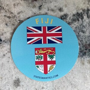 Könnte beinhalten: Ein runder blauer Aufkleber mit dem Wort "FIJI" in gelben Buchstaben. Der Aufkleber zeigt die Flagge von Fidschi mit einem Union Jack in der oberen linken Ecke und dem Wappen von Fidschi in der unteren Mitte. Der Text "STATESMANTIES.COM" ist unten aufgedruckt.
