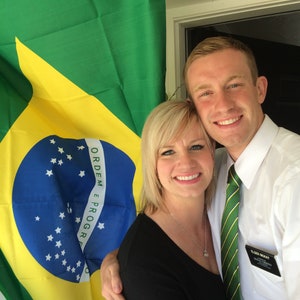 May include: A couple poses in front of a Brazilian flag. The man is wearing a white shirt with a green tie and a name tag that says "Elder McKay". The woman is wearing a black shirt and a silver necklace. The flag has the words "Ordem e Progresso" written on it.
