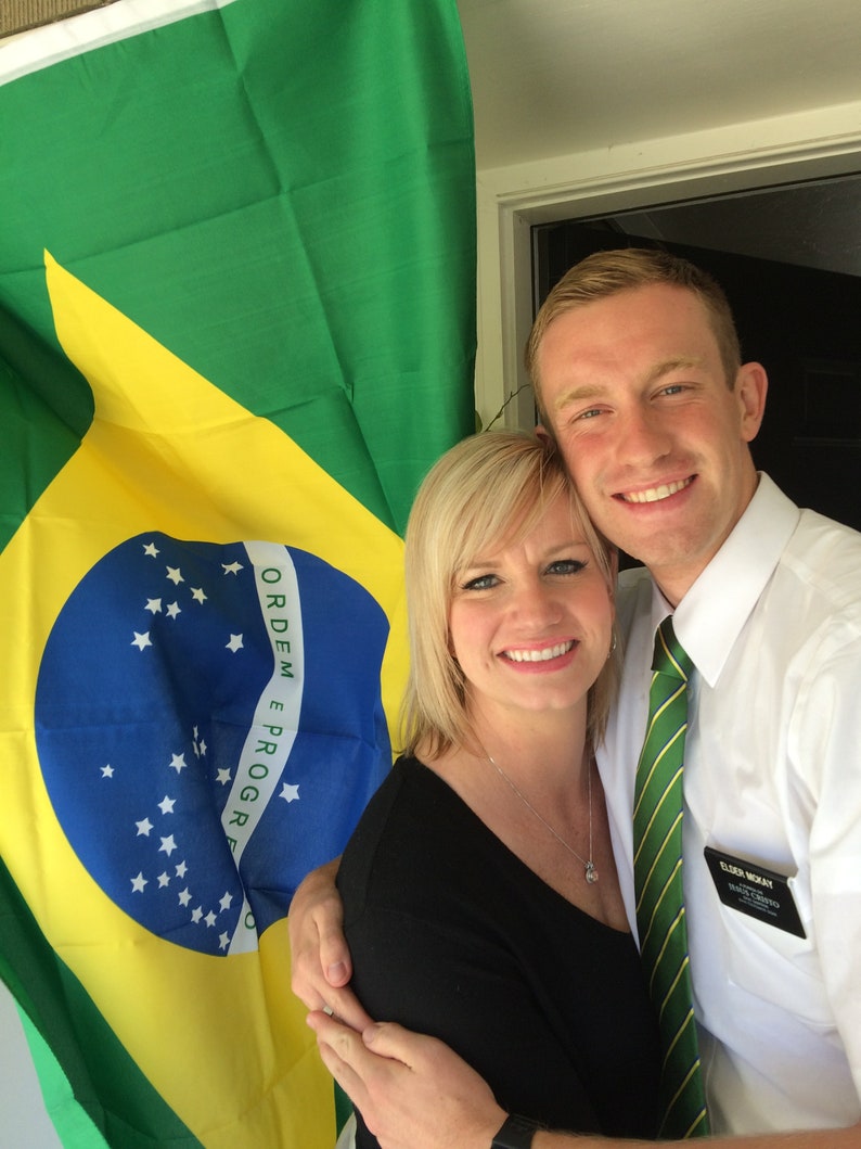May include: A couple poses in front of a Brazilian flag. The man is wearing a white shirt with a green tie and a name tag that says "Elder McKay". The woman is wearing a black shirt and a silver necklace. The flag has the words "Ordem e Progresso" written on it.