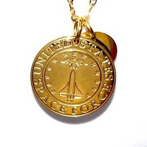 Op de afbeelding: Gouden ketting met een ronde hanger met het logo van de United States Space Force. Het logo bevat een spaceshuttle, een adelaar en sterren.