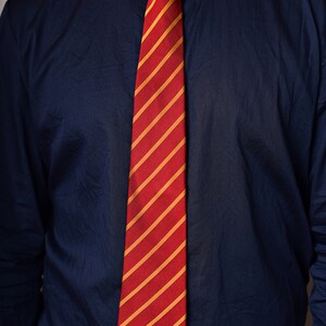 May include: A red and gold striped tie with a small gold emblem. The tie is worn with a dark blue button-down shirt.