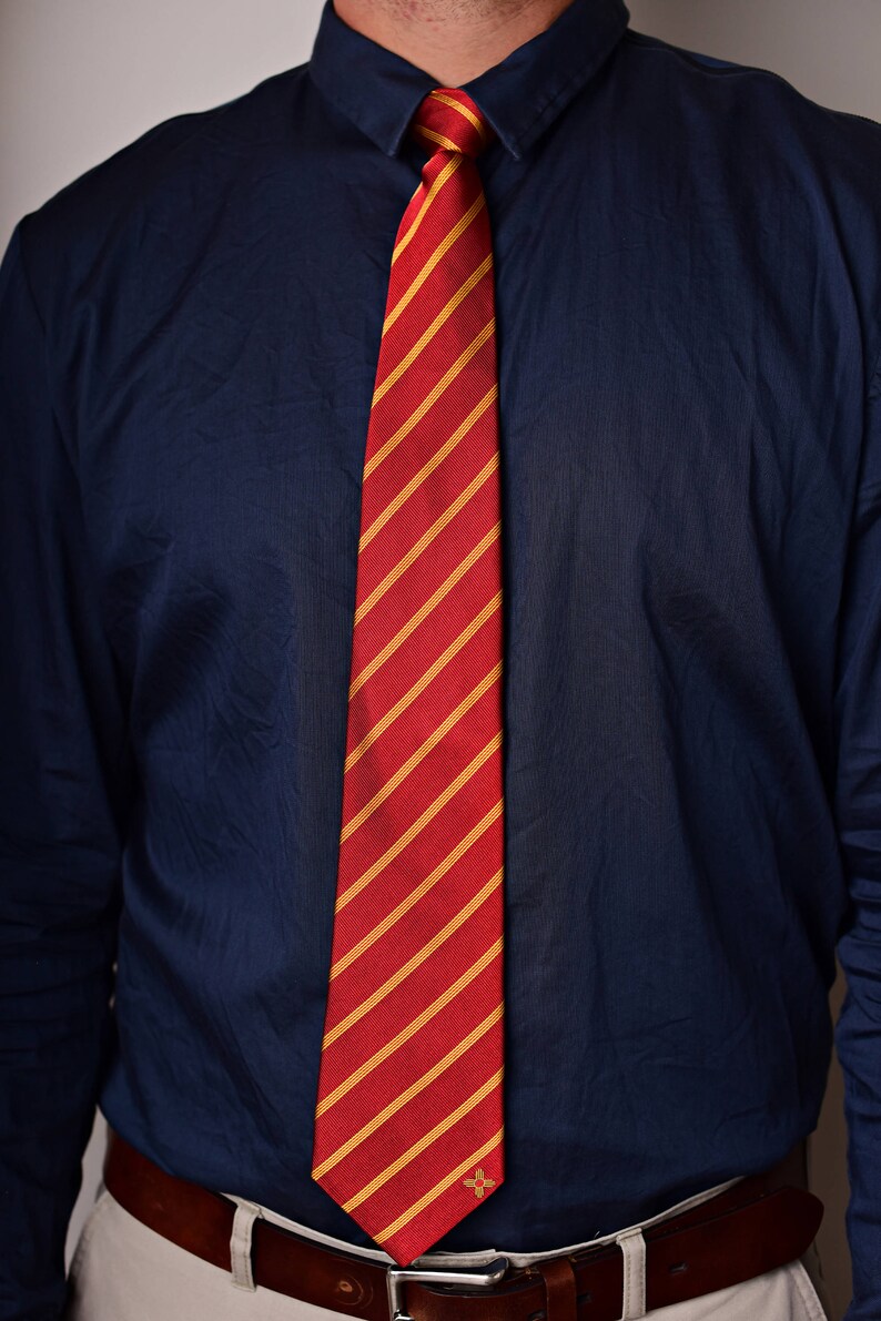 May include: A red and gold striped tie with a small gold emblem. The tie is worn with a dark blue button-down shirt.
