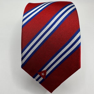 Op de afbeelding: Een rode stropdas met witte en blauwe diagonale strepen. De stropdas heeft een klein geborduurd Cubaans vlaggetje aan de onderkant.