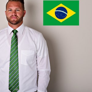 May include: A man wearing a white dress shirt with a green and white striped tie featuring a small Brazilian flag. The tie has a green, white, and blue striped pattern. The man is standing in front of a white wall with a Brazilian flag on it. The flag is green, yellow, and blue with a blue circle in the center. The text on the flag reads "Ordem e Progresso".