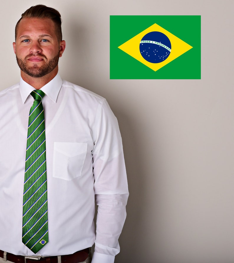 May include: A man wearing a white dress shirt with a green and white striped tie featuring a small Brazilian flag. The tie has a green, white, and blue striped pattern. The man is standing in front of a white wall with a Brazilian flag on it. The flag is green, yellow, and blue with a blue circle in the center. The text on the flag reads "Ordem e Progresso".