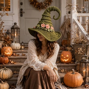 May include: A pointed olive green crochet witch hat featuring a curled tip and a decorative band of small pink and light green fabric flowers around the base. The hat has a wide, textured brim and is crafted with a detailed, ribbed stitch pattern.
