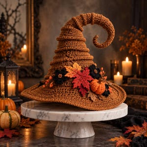 Puede incluir: Un sombrero de bruja de ganchillo marrón con punta curvada, decorado con un grupo de hojas de otoño de tela en tonos naranja, rojo y amarillo, pequeñas flores de ganchillo negras y una calabaza de ganchillo naranja en miniatura.