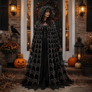 May include: A floor-length black hooded cloak featuring an intricate web-like lace pattern with metallic gold accents. The garment is worn over a solid black dress with a corset-style bodice, creating a layered appearance with long, flowing fabric.