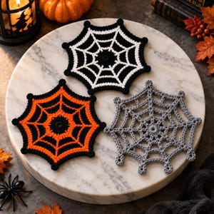 May include: Three crocheted spider web coasters in a hexagonal shape. One coaster is black with white web detailing, one is orange with black web detailing, and one is solid grey. Each coaster features a central circular point with radiating lines.