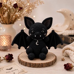 May include: A black crocheted bat plush toy with large dark eyes, two small white fangs, and a silver crescent moon charm necklace. The bat has pointed ears and outstretched wings, sitting upright on a circular wooden slice base.