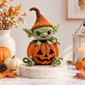 May include: A crocheted green goblin figure with large black eyes and a pointed orange hat, sitting inside a round orange crocheted pumpkin with a black stitched jack-o'-lantern face and green curly vines extending from the base.