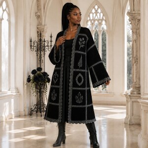 Può includere: Un lungo cardigan nero aperto sul davanti con un design patchwork a pannelli quadrati. Ogni pannello è bordato da cuciture grigie e presenta motivi ricamati tra cui fiori, felci, cornici ornate e gruppi di cristalli. Maniche ampie.