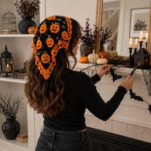 May include: A black crochet headscarf featuring an orange pumpkin pattern and a scalloped orange border. The triangular headscarf is worn over long brown hair, showcasing a detailed knit texture with repeating jack-o'-lantern motifs throughout.