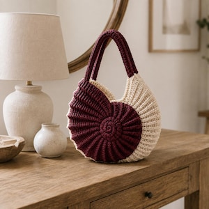 May include: A circular crochet handbag featuring a nautilus shell design. The bag is constructed with two distinct colour sections, one in deep burgundy and the other in cream, with a spiral centre and ribbed texture throughout the entire surface.